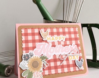 May include: A pink Mother's Day card with a gingham pattern and floral embellishments. The card features the words "Happy Mother's Day" in a cursive font. Decorative elements include a sunflower, chickens, and a bow.