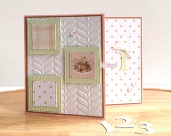 May include: Two handmade greeting cards. One card has a white embossed background with three framed squares. The other card is pink with gold polka dots and a number one. Wooden numbers 1, 2, and 3 are on the wooden surface.
