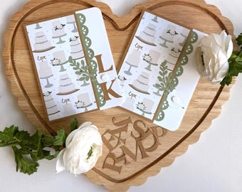 May include: Two cards with a cake pattern and the word "Love" are displayed on a wooden heart-shaped tray. The cards have green lace and floral accents. The letters "L" and "K" are visible on one card. White flowers and greenery are also present. The tray is heart-shaped.