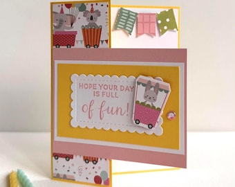 May include: A handmade birthday card with a yellow and pink design. The card features illustrations of animals in carts, decorative banners, and the text "HOPE YOUR DAY IS FULL of fun!" Several colourful birthday candles are in the foreground.