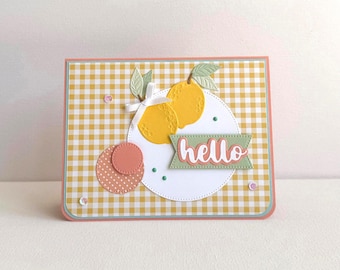 May include: A handmade greeting card with a yellow and white gingham pattern. The card features a white circle with two yellow lemon cutouts, green leaves, and a banner that says "hello". Additional pink and peach circles are included.