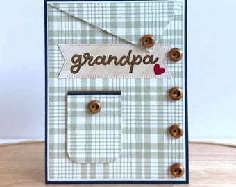 May include: A handmade greeting card with a green and white plaid pattern. The card features the word "grandpa" on a banner, a red heart, and several brown buttons. The card has a navy blue border.