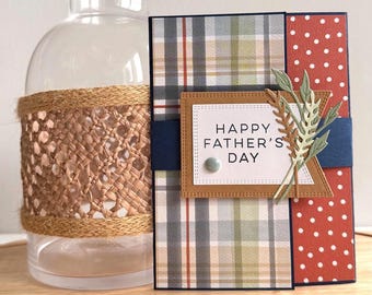 May include: A Father's Day card with a plaid design, a red polka dot panel, and a navy blue ribbon. The card reads "HAPPY FATHER'S DAY" and is adorned with wheat stalks. A clear glass bottle with a woven raffia sleeve is in the background.