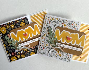 May include: Two handmade Mother's Day cards. Each card has a brown label with the word "MOM" in yellow, a red heart, and the phrase "GRATEFUL FOR YOU." The cards feature floral and striped patterns in various colours.