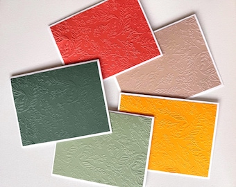 May include: A selection of embossed greeting cards in shades of red, green, beige, yellow, and pale green. Each card displays a floral design and is framed with a white border. The cards are arranged on a white surface.