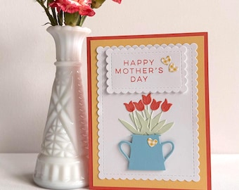 May include: A Mother's Day card featuring a blue jug with red tulips, set against a yellow and red background. The card reads "HAPPY MOTHER'S DAY". A white vase with red flowers is positioned to the left of the card.
