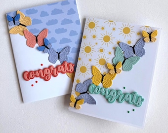 May include: Two greeting cards with butterfly and "congrats" designs. One card has a blue sky and cloud pattern, the other has a yellow sun pattern. The cards feature colourful butterflies in yellow, blue, and coral.