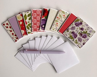 May include: A selection of patterned stationery cards with white envelopes. The cards display floral and geometric designs in red, purple, and green. A lavender pen is placed on the envelopes. The cards are ideal for sending notes.