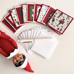 May include: A set of square holiday greeting cards with red borders and various Christmas-themed patterns including elves, snowflakes, and string lights, accompanied by a stack of white envelopes, a white pen, and a plush elf doll with a red hat.