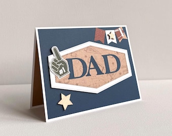 May include: A handmade Father's Day card with a navy blue background and a white border. The card features a brown and white hexagon with the word "DAD" in large letters. A foam finger and wooden star add decorative elements.
