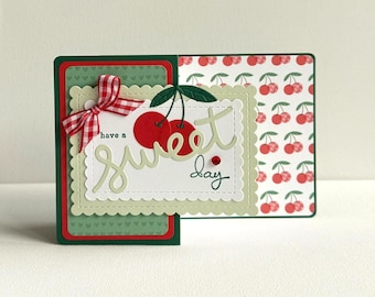 May include: A handmade greeting card with a cherry theme. The card features a red and green colour scheme, a gingham bow, and the words "have a sweet day". The inside of the card has a cherry pattern.