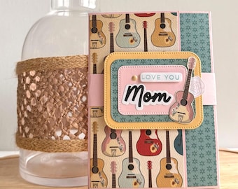 May include: A handmade greeting card with a guitar pattern and the words "Love You Mom." The card features a pink and teal colour scheme with a decorative guitar image. A clear glass bottle with a woven brown wrap is in the background.