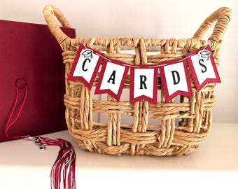May include: A woven wicker basket with handles, decorated with a banner that reads "CARDS" in black letters on white pennants with red borders. A graduation cap and tassel are visible on the left.