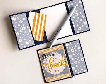 May include: A navy blue card holder with a daisy pattern and a yellow and white striped card. A light blue pen rests on the open card holder. A small card with the word "Thanks" in gold script is also present.