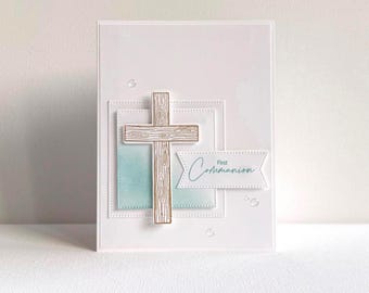 May include: A white card featuring a wooden cross and the words "First Communion" in light blue script. The cross is made of light brown wood and is centered on the card. The card has a layered design with a light blue watercolor background.