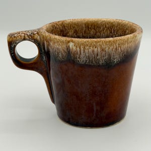 Pode incluir: Uma caneca de cerâmica com acabamento de esmalte escorrido em dois tons. A parte superior apresenta um padrão salpicado de branco creme e castanho claro que transita para um esmalte castanho escuro sólido na base e na asa circular.
