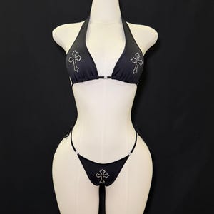May include: Black halter bikini set with rhinestone cross embellishments. The top features cross designs on each cup, and the bottoms have a matching cross. The bikini is displayed on a mannequin against a black backdrop.