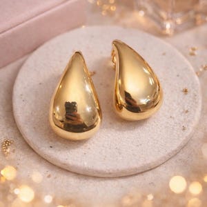 May include: Two gold teardrop earrings with a high-shine finish. The earrings are displayed on a circular, pale stone surface. The background is softly blurred, featuring pink and gold tones, with a hint of a perfume bottle.
