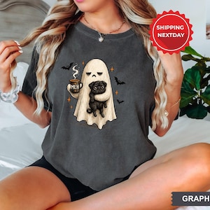May include: A graphite gray t-shirt featuring a graphic of a ghost with a bored expression holding a steaming mug labeled MEH and a black pug. The design includes small orange stars and black bats surrounding the central ghost and dog illustration.