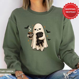 May include: A military green crewneck sweatshirt featuring a graphic of a white ghost holding a brown mug with the text MEH and a black pug. The design includes small orange stars and black bats surrounding the ghost on the front of the garment.