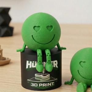 Könnte beinhalten: Zwei grüne, kugelförmige Figuren mit herzförmigen Augen und lächelnden Gesichtern. Eine sitzt auf einem schwarzen Zylinder mit dem Text "HÜ R 3D PRINT". Die andere sitzt auf einer Holzoberfläche. 3D-Druckzubehör im Hintergrund.