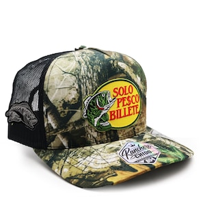 May include: A camouflage trucker hat with a black mesh back and a front patch featuring a green fish and the text SOLO PESCO BILLETE in red and yellow. A smaller fish patch is on the side, and a Ranchero Chido sticker is on the flat brim.