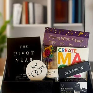May include: A black gift box filled with various items, including a book titled "The Pivot Year," a round "Self Love" sign, and a "Flying Wish Paper" kit. Other items include a colorful floral arrangement and a "Create" book.
