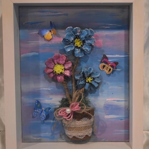 May include: A framed artwork featuring a floral arrangement with blue and pink flowers, crafted from pinecones. Butterflies in various colors are scattered around the flowers. The background is painted with blue, pink, and white hues.
