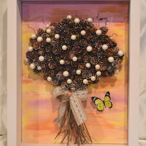 May include: A framed artwork featuring a bouquet made of pinecones and adorned with white pearl-like beads. The bouquet is tied with a beige ribbon and rests on a painted background with a butterfly. The frame is white.