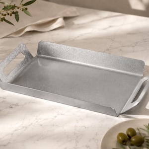 May include: A rectangular silver-colored metal tray with a textured, galvanized finish. The tray features two integrated handles on the shorter sides and raised edges along the perimeter, providing a flat, open surface for holding various items.