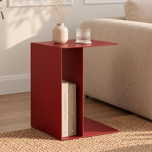 May include: A deep red metal C-shaped side table featuring a flat top surface and a vertical storage compartment. The interior shelf holds two books with spines labelled The Kinfolk and Atelier AM, while the top surface holds a small vase and glass.