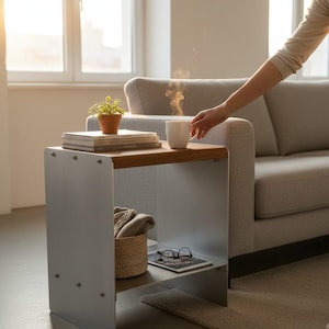 Puede incluir: Una mesa auxiliar con una superficie de madera clara y una estructura de metal plateado de dos niveles. La parte superior sostiene una pila de libros, una pequeña planta en maceta y una taza blanca. El estante inferior contiene una cesta tejida y gafas.