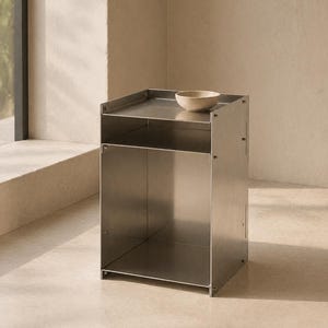 May include: A rectangular stainless steel side table with a smooth metallic finish, featuring an open storage compartment and a flat top surface. A small, plain beige ceramic bowl is placed on the top shelf of this modular metal furniture piece.