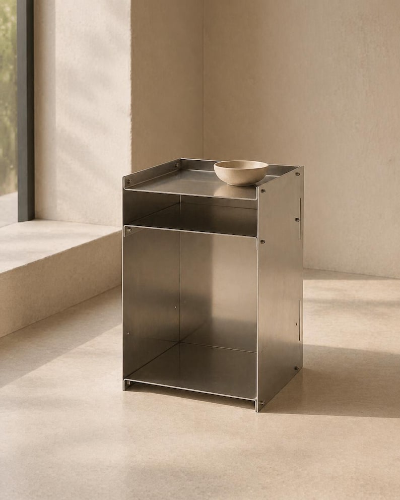 May include: A rectangular stainless steel side table with a smooth metallic finish, featuring an open storage compartment and a flat top surface. A small, plain beige ceramic bowl is placed on the top shelf of this modular metal furniture piece.