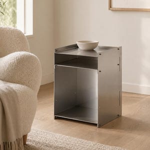 May include: A brushed metal side table with a rectangular frame, featuring an open storage compartment and a flat top surface. A small, off-white ceramic bowl sits on the top shelf of the table, which is constructed from joined metal panels.