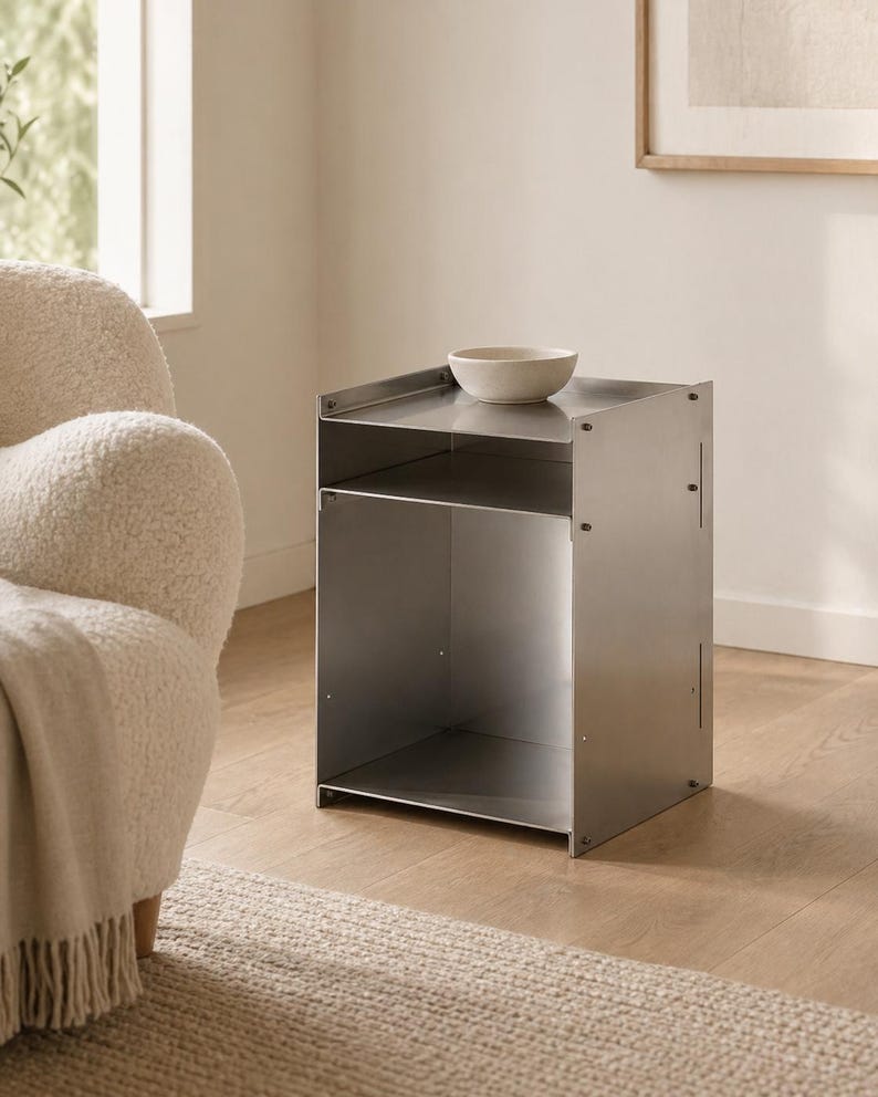 May include: A brushed metal side table with a rectangular frame, featuring an open storage compartment and a flat top surface. A small, off-white ceramic bowl sits on the top shelf of the table, which is constructed from joined metal panels.