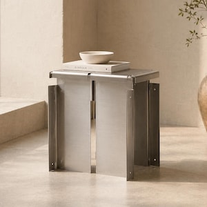 Könnte beinhalten: Ein moderner Beistelltisch aus gebürstetem Metall mit rechteckiger Oberseite und einem einzigartigen, offenen Design. Eine kleine, weiße Schale und ein Buch liegen obenauf. Der Tisch ist etwa 46 cm hoch.