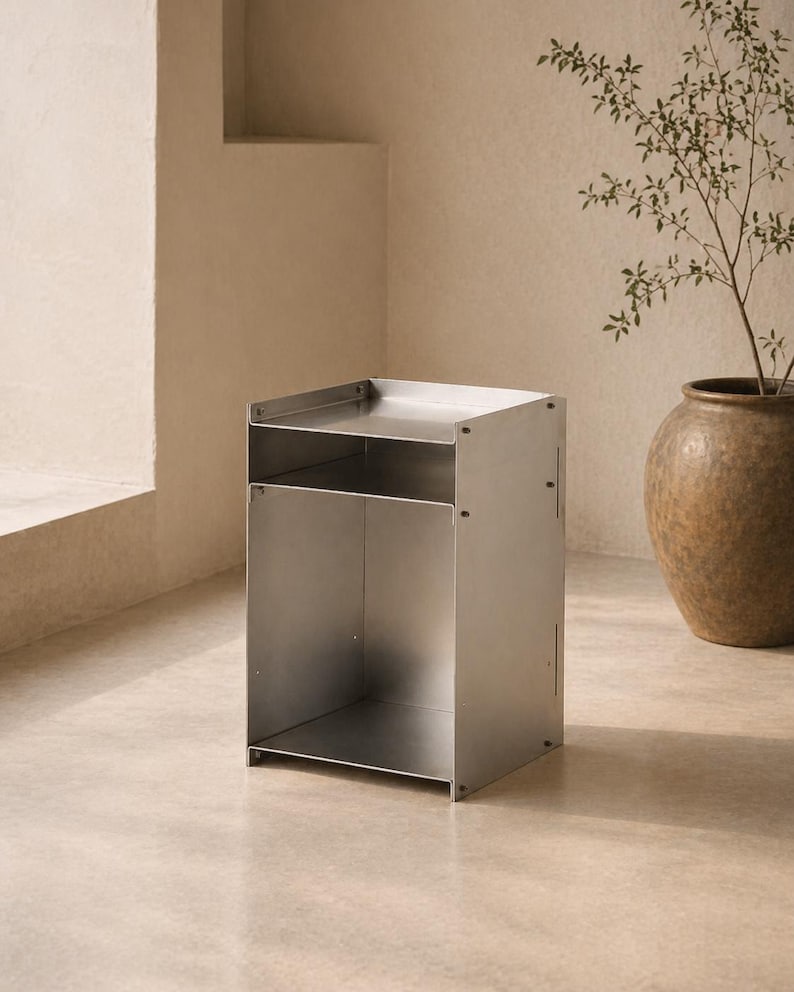 May include: A rectangular metal side table constructed from brushed silver-toned panels. The piece features a flat top surface, an open middle shelf, and a larger open storage compartment at the base, all held together by visible metal fasteners.