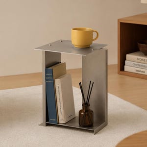 Puede incluir: Una mesa auxiliar de metal cepillado con una superficie rectangular y un estante inferior. La mesa sostiene una taza de cerámica amarilla en la parte superior, mientras que el estante inferior contiene dos libros con lomos azul y blanco y una botella de vidrio marrón con difusores de varillas.