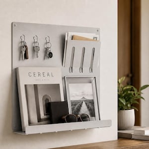 May include: A silver metal wall organiser with key hooks, a mail slot, and a shelf. It holds keys, magazines, a framed photo, a wallet, and sunglasses. The magazine cover reads "CEREAL TRAVEL & STYLE."