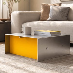 May include: A low-profile coffee table constructed from brushed stainless steel with a vibrant yellow interior panel. The table features a flat rectangular top surface, two small metal fasteners on the front face, and a mug and two books placed on top.