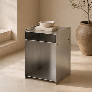 May include: A silver metal side table with a shelf and open compartment. A beige bowl and a book rest on top. The table is rectangular and has visible screws. A large, brown ceramic pot with a plant is in the background.