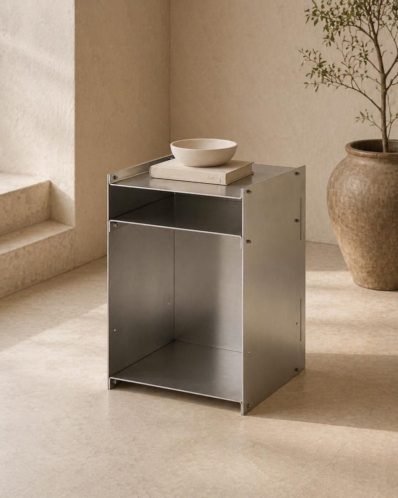 May include: A rectangular metal side table with a brushed silver finish, featuring an open storage compartment and a flat top surface. A beige book and a small, shallow beige bowl are placed on the top of the metal structure.