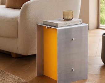 May include: A rectangular side table featuring a brushed silver metal exterior and a bright yellow interior panel. The table has two small silver knobs on the front face. A book titled KINFOLK and a ribbed amber glass candle sit on the top surface.