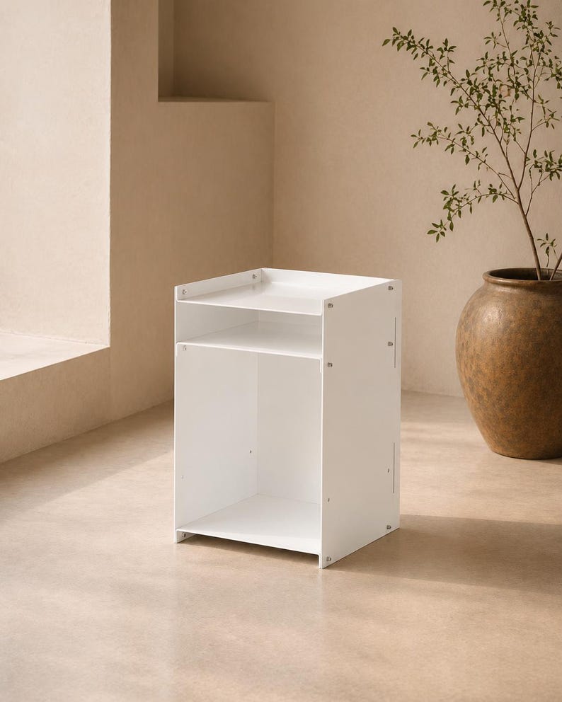 May include: A white bedside table with a rectangular frame, featuring an open top shelf and a larger open bottom storage compartment. The unit is constructed from flat panels joined by visible metal screws on the side panels, creating a clean, functional storage piece.
