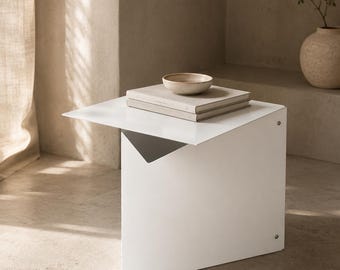 May include: A white metal side table with a flat top and a triangular base structure. Two stacked books and a small, shallow ceramic bowl are placed on the surface. The table features visible metal fasteners on the side panel and a clean finish.