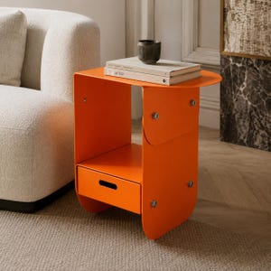 Può includere: Un tavolino arancione con un cassetto e un piano arrotondato. Il tavolo ha un design moderno ed è realizzato in metallo. Una piccola tazza di ceramica scura e due libri sono sopra.