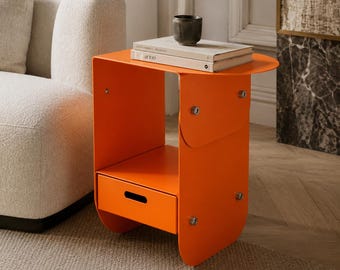 May include: An orange side table with a drawer and a rounded top. The table has a modern design and is made of metal. A small, dark-colored ceramic cup and two books are on top of the table.