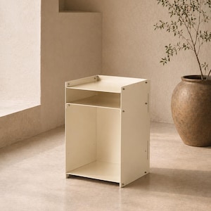 May include: A cream-colored metal side table featuring a rectangular frame with a flat top surface, an open middle shelf, and a larger open storage compartment at the base. The structure is held together by visible silver-toned screws on the sides.
