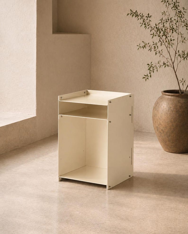 May include: A cream-colored metal side table featuring a rectangular frame with a flat top surface, an open middle shelf, and a larger open storage compartment at the base. The structure is held together by visible silver-toned screws on the sides.